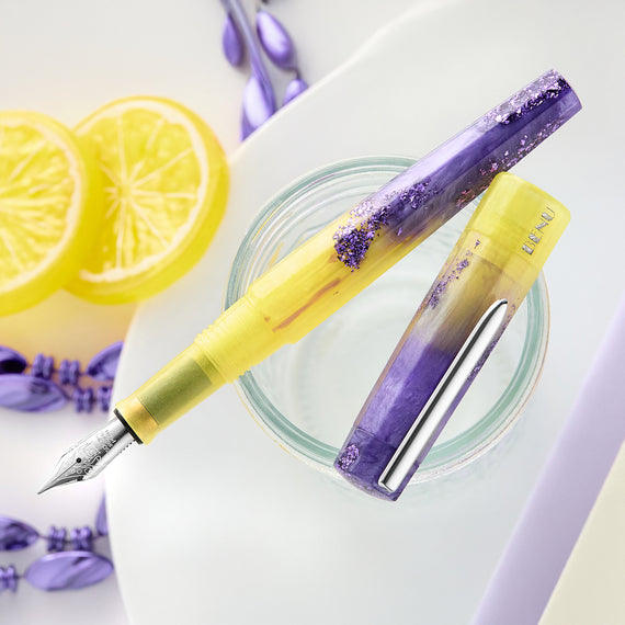Yellow and purple fountain pen with decorative cap on a white surface with lemon slices and lavender flowers.