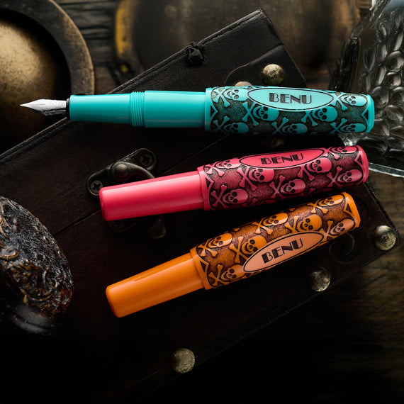 Three BENU pens in turquoise, pink, and orange on a dark surface with decorative elements.