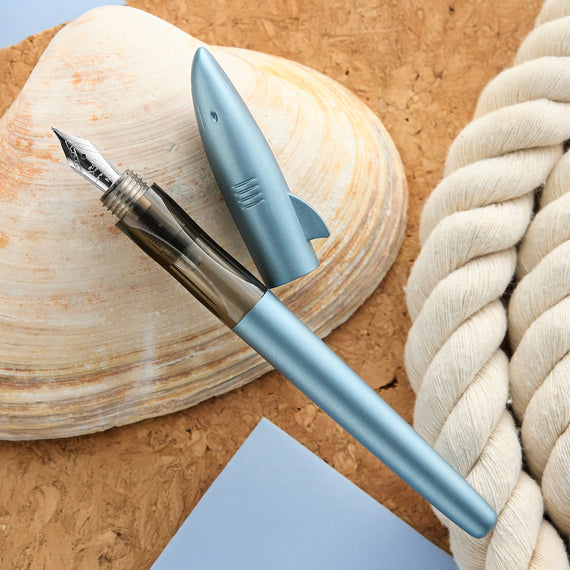 Blue shark-shaped pen on a wooden surface with a shell and rope.