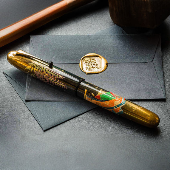 Decorative pen with artistic design on a dark surface next to a black envelope with a gold seal.
