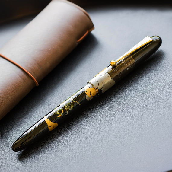 Fountain pen with floral design on a dark surface next to a brown leather case.