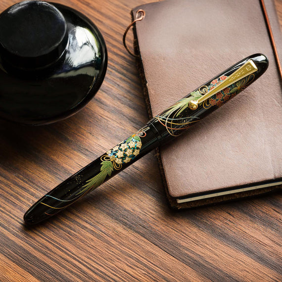 Decorative pen with floral design on a wooden surface next to a notebook and black inkwell.