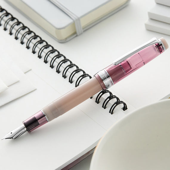 Pink and silver fountain pen on a white surface with a notebook and cup in the background