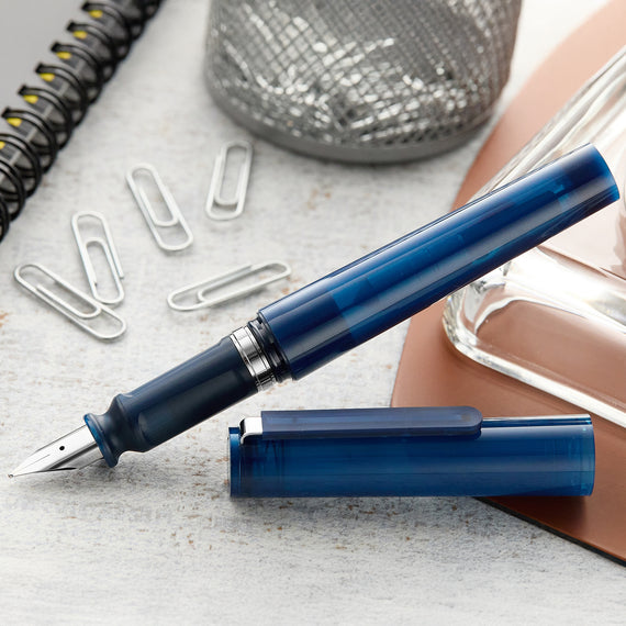 Blue fountain pen on a light surface with paper clips and a glass container.
