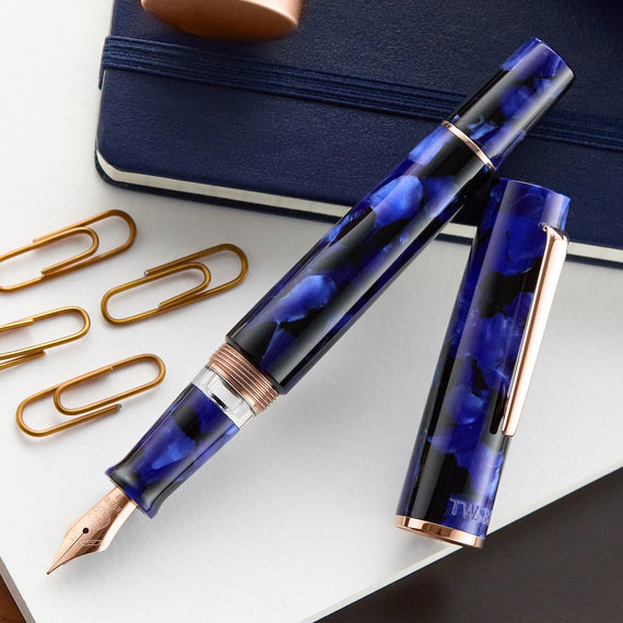 Blue patterned fountain pen with rose gold accents on a white surface with a notebook and paper clips.